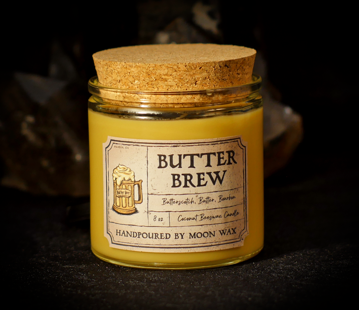 Butter Brew Candle in a clear corked jar with honey colored coconut beeswax and apothecary inspired label on a dark background. The label reads Butterscotch, Butter, Bourbon, 8 ounces, and hand poured by Moon Wax.
