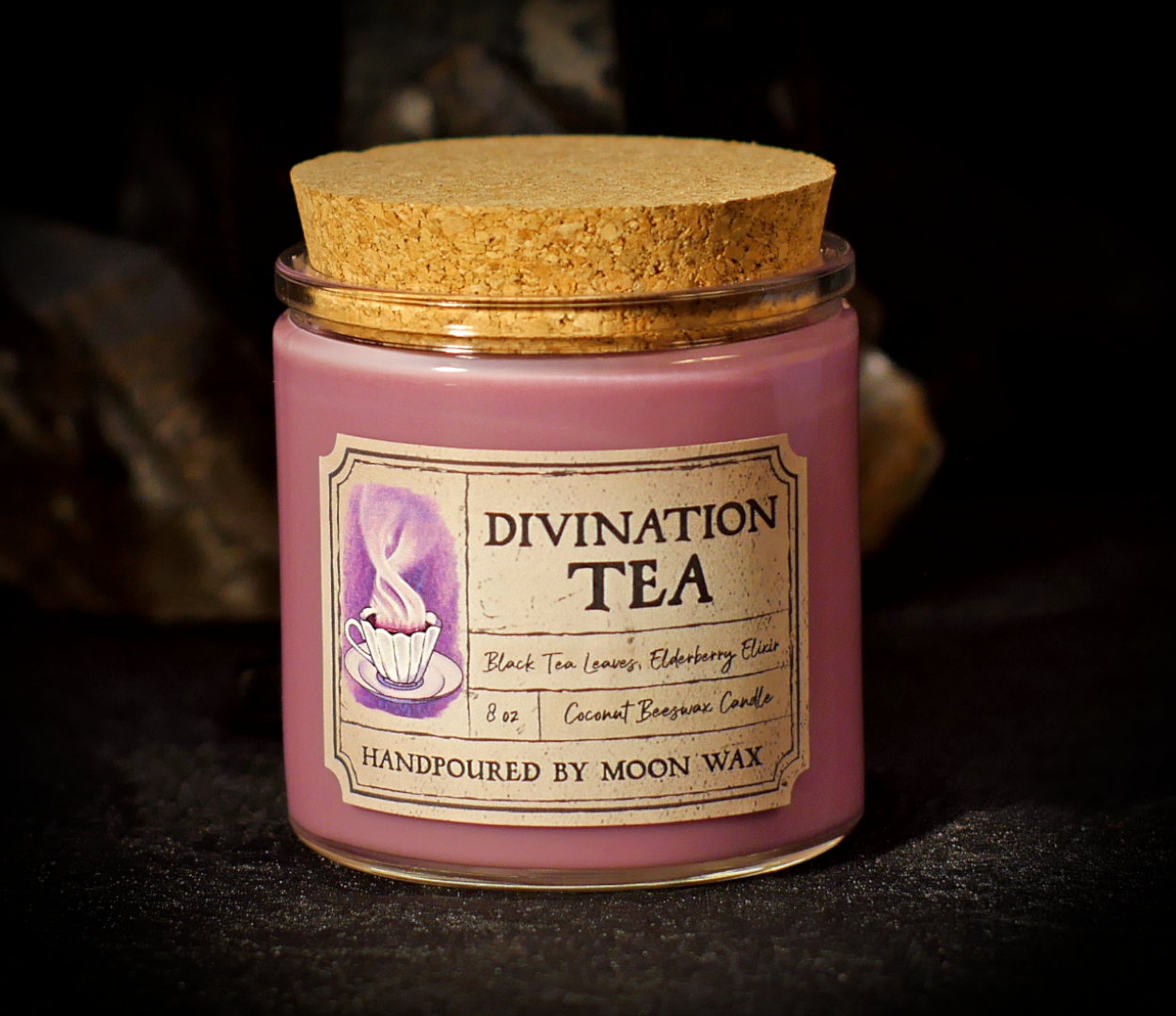 Divination Tea Candle in a clear corked jar with magenta colored coconut beeswax and apothecary inspired label on a dark background. The label reads Black Tea Leaves, Elderberry Elixir, 8 ounces, and hand poured by Moon Wax.