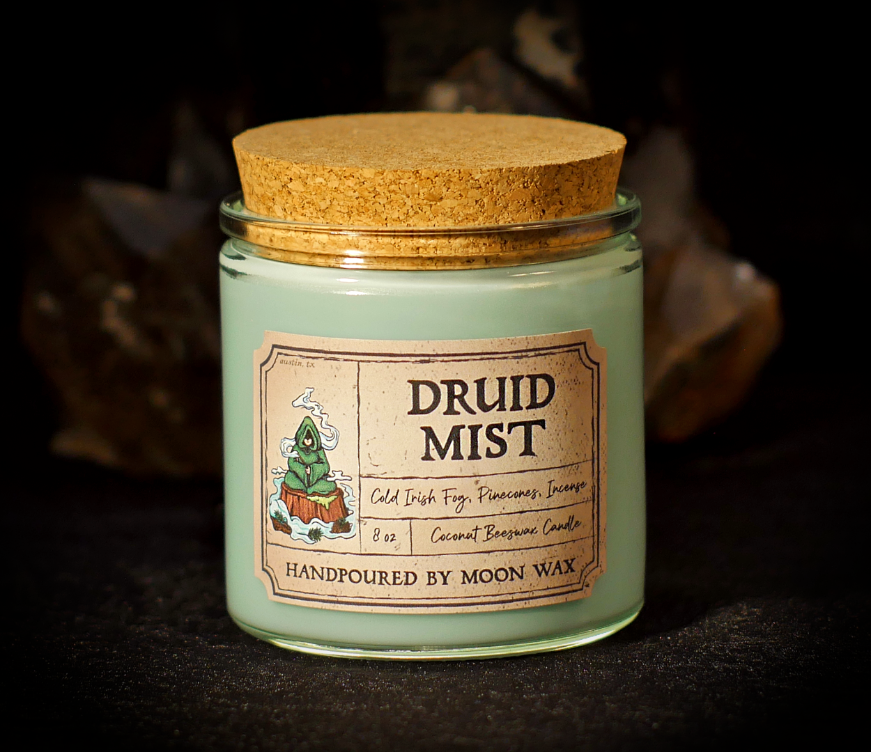 Druid Candle in a clear corked jar with green colored coconut beeswax and apothecary inspired label on a dark background. The label reads Cold Irish Fog, Pinecones, Incense, 8 ounces, and hand poured by Moon Wax.