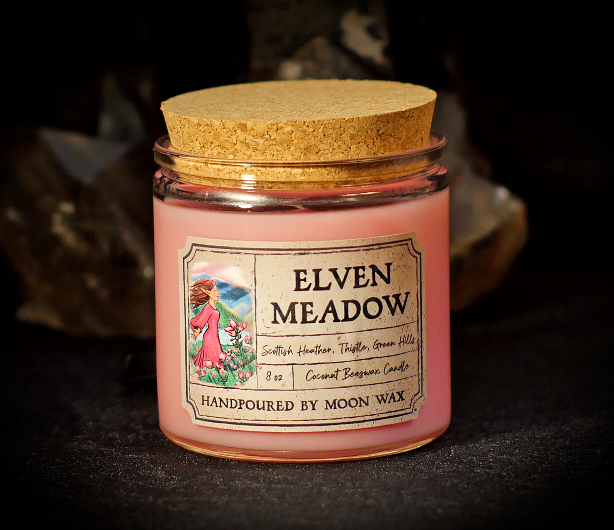 Elven Meadow Candle in a clear corked jar with pink colored coconut beeswax and apothecary inspired label on a dark background. The label reads Scottish Heather, Thistle, Green Hills, 8 ounces, and hand poured by Moon Wax.