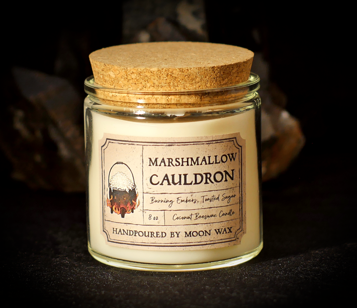 Marshmallow Cauldron Candle in a clear corked jar with teal-blue colored coconut beeswax and apothecary inspired label on a dark background. The label reads Burning Embers, Toasted Sugar, 8 ounces, and hand poured by Moon Wax.