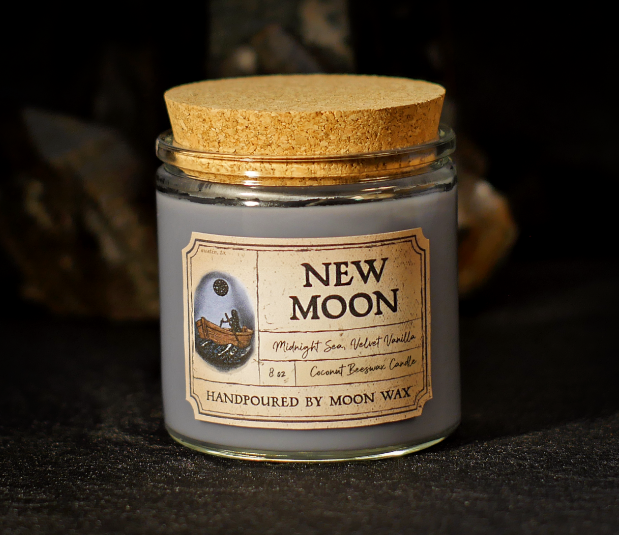 New Moon Candle in a clear corked jar with dark-gray colored coconut beeswax and apothecary inspired label on a dark background. The label reads Midnight Sea, Velvet Vanilla, 8 ounces, and hand poured by Moon Wax.