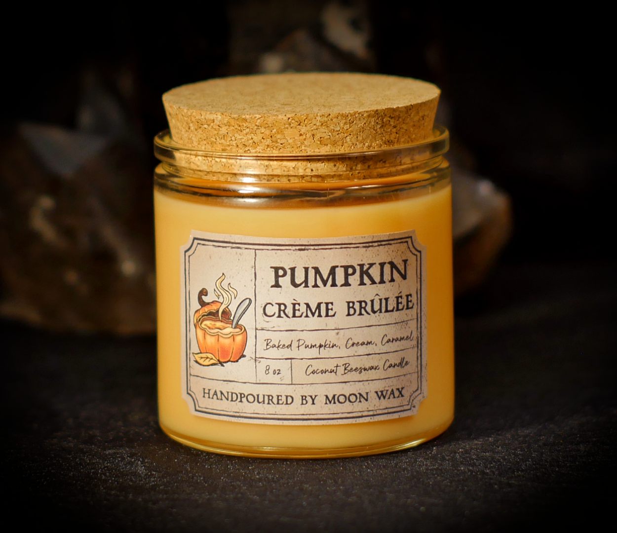 Pumpkin Creme Brulé Candle in a clear corked jar with orange colored coconut beeswax and apothecary inspired label on a dark background. The label reads Baked Pumpkin, Cream, Caramel, 8 ounces, and hand poured by Moon Wax.
