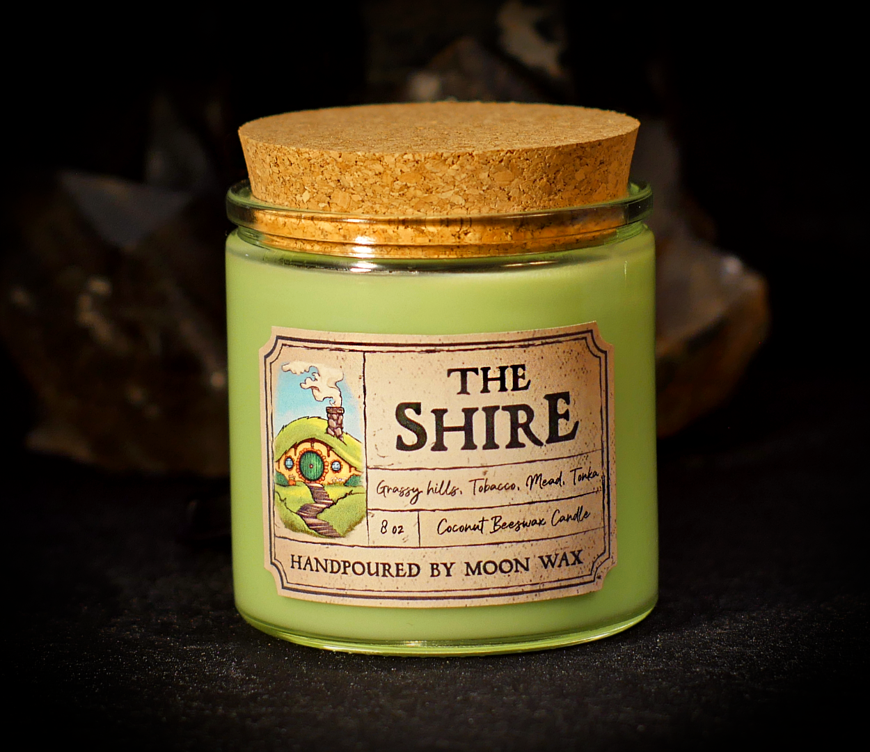 The Shire Candle in a clear corked jar with lime-green colored coconut beeswax and apothecary inspired label on a dark background. The label reads Grassy Hills, Tobacco, Mead, Tonka, 8 ounces, and hand poured by Moon Wax.